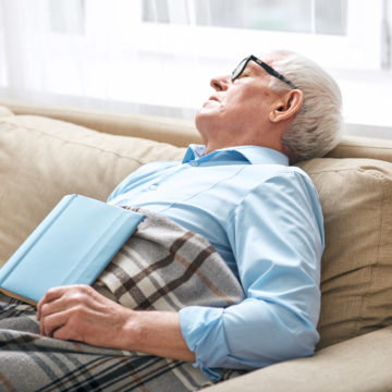 Tired senior man covered with plaid napping on couch with open book CASI CINCO DE CADA DIEZ ESPAÑOLES DUERMEN LA SIESTA A DIARIO DESDE QUE EMPEZÓ LA PANDEMIA